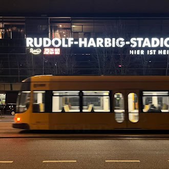 Nachtaufnahme des Rudolf-Harbig-Stadions in Dresden mit leuchtender Schrift „Rudolf-Harbig-Stadion Hier ist Heimat“. Im Vordergrund fährt eine gelbe DVB-Straßenbahn in Bewegungsunschärfe auf der Straße vorbei.