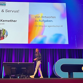 Sarah Kemether von OpenAI geht über eine violett beleuchtete Konferenzbühne. Auf einer Leinwand hinter ihr ist ihr Vortragstitel „Von Antworten zu Aufgaben. Der Weg zur agentischen KI“ sowie ihr Porträtfoto zu sehen.