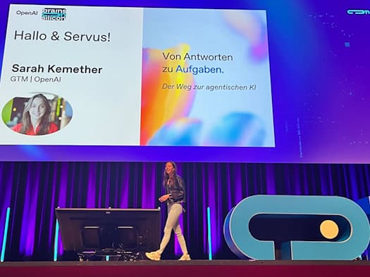 Sarah Kemether of OpenAI walks across a conference stage illuminated in purple. On a screen behind her, the title of her presentation, “From Answers to Tasks: The Path to Agent-Based AI,” and her portrait photo are displayed.