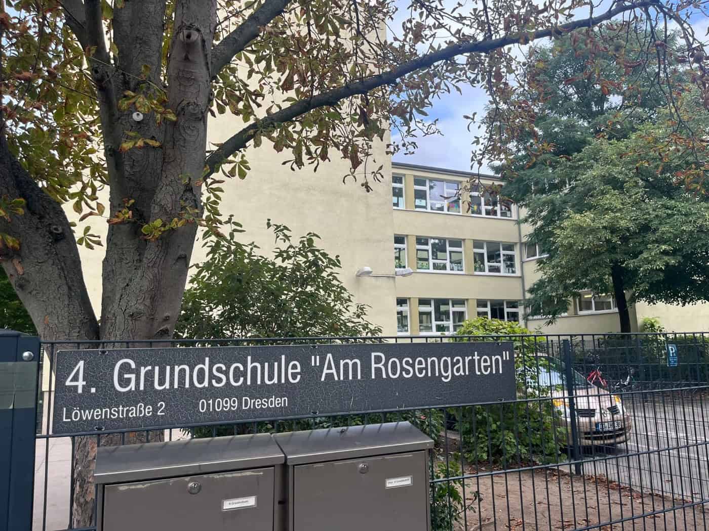 Nahaufnahme des dunklen Eingangsschildes der 4. Grundschule „Am Rosengarten“ an einem Metallzaun in der Löwenstraße 2 in Dresden. Dahinter befindet sich das mehrstöckige Schulgebäude hinter den Ästen eines großen Baumes.