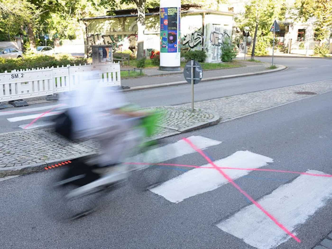 Ein weiß auf den Asphalt gemalter Zebrastreifen auf einer Straße, der mit neonroten Strichen durchgekreuzt wurde. Ein Radfahrer fährt verschwommen im Vordergrund vorbei. Im Hintergrund stehen weiße Absperrgitter auf einer Mittelinsel.