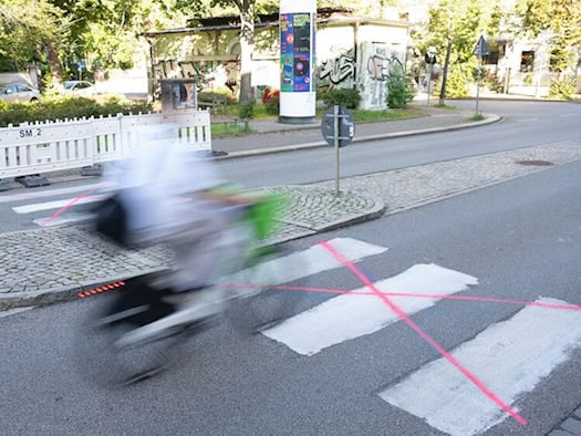 Ein weiß auf den Asphalt gemalter Zebrastreifen auf einer Straße, der mit neonroten Strichen durchgekreuzt wurde. Ein Radfahrer fährt verschwommen im Vordergrund vorbei. Im Hintergrund stehen weiße Absperrgitter auf einer Mittelinsel.