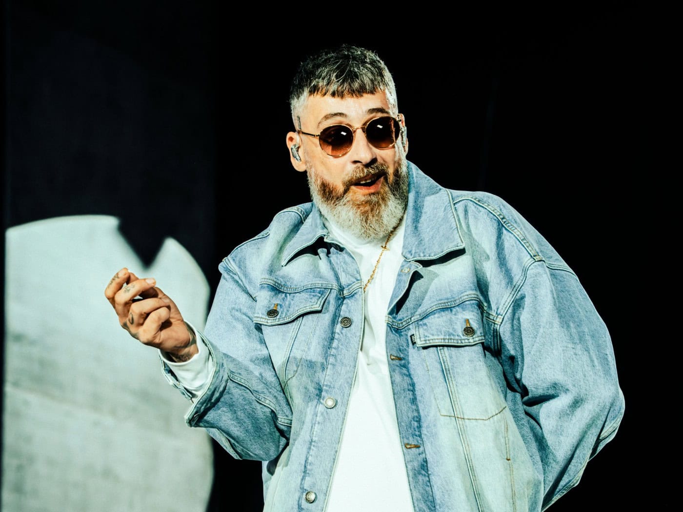 Der Berliner Rapper Sido steht mit ergrautem Vollbart, Sonnenbrille und heller Jeanskleidung (Jeansjacke und weite Jeanshose) auf einer Konzertbühne. Er hält eine Hand gehoben und gestikuliert während eines Auftritts.