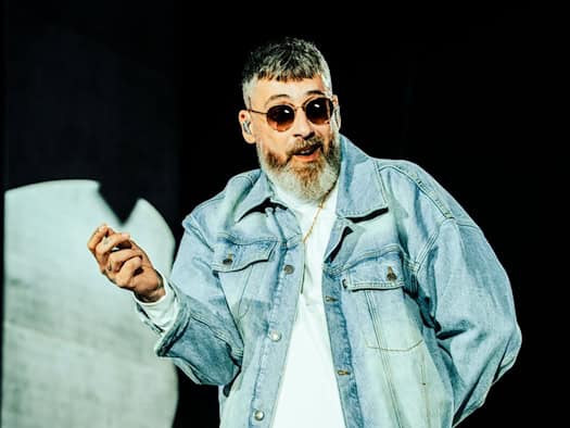 Der Berliner Rapper Sido steht mit ergrautem Vollbart, Sonnenbrille und heller Jeanskleidung (Jeansjacke und weite Jeanshose) auf einer Konzertbühne. Er hält eine Hand gehoben und gestikuliert während eines Auftritts.