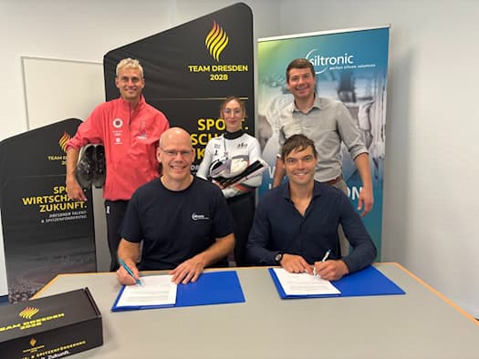 Zwei Männer sitzen an einem Tisch und unterzeichnen Verträge. Dahinter stehen Leichtathlet Karl Bebendorf mit Laufschuhen, Shorttrackerin Lisa Eckstein mit Schlittschuhen und ein weiterer Mann vor Bannern von Siltronic und Team Dresden 2028.