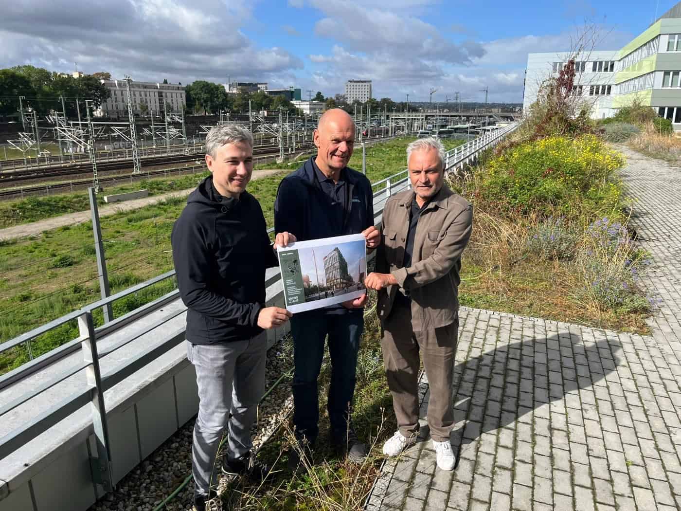 Drei Männer stehen auf einer Dachterrasse und halten eine Visualisierung des geplanten Simmel Terminals, dahinter Bahngleise und der Dresdner Hauptbahnhof.