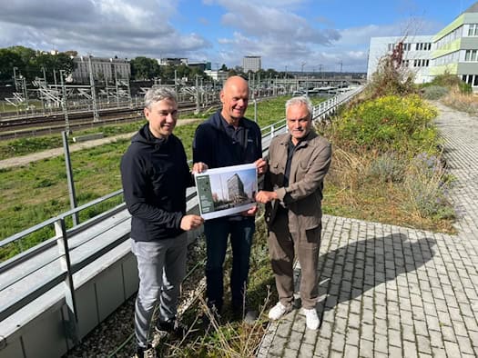 Drei Männer stehen auf einer Dachterrasse und halten eine Visualisierung des geplanten Simmel Terminals, dahinter Bahngleise und der Dresdner Hauptbahnhof.