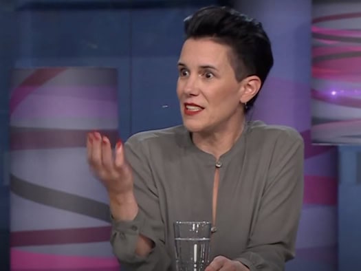 Simone Meyer-Götz mit kurzen dunklen Haaren und grauem Oberteil sitzt in einem Fernsehstudio. Sie blickt konzentriert zur Seite und gestikuliert lebhaft mit der rechten Hand mit rot lackierten Fingernägeln, vor ihr steht ein Glas Wasser auf dem Tisch.