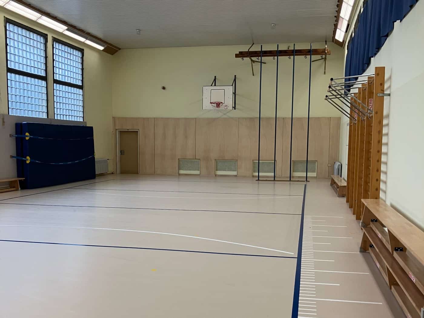 Blick in den hellen Innenraum einer sanierten Turnhalle mit hellem Linoleumboden und blau-weißen Spielfeldlinien. An den Wänden stehen hölzerne Sprossenwände und Bänke, im Hintergrund sind Kletterseile und ein Basketballbrett zu sehen.