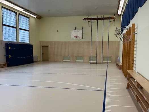 Blick in den hellen Innenraum einer sanierten Turnhalle mit hellem Linoleumboden und blau-weißen Spielfeldlinien. An den Wänden stehen hölzerne Sprossenwände und Bänke, im Hintergrund sind Kletterseile und ein Basketballbrett zu sehen.