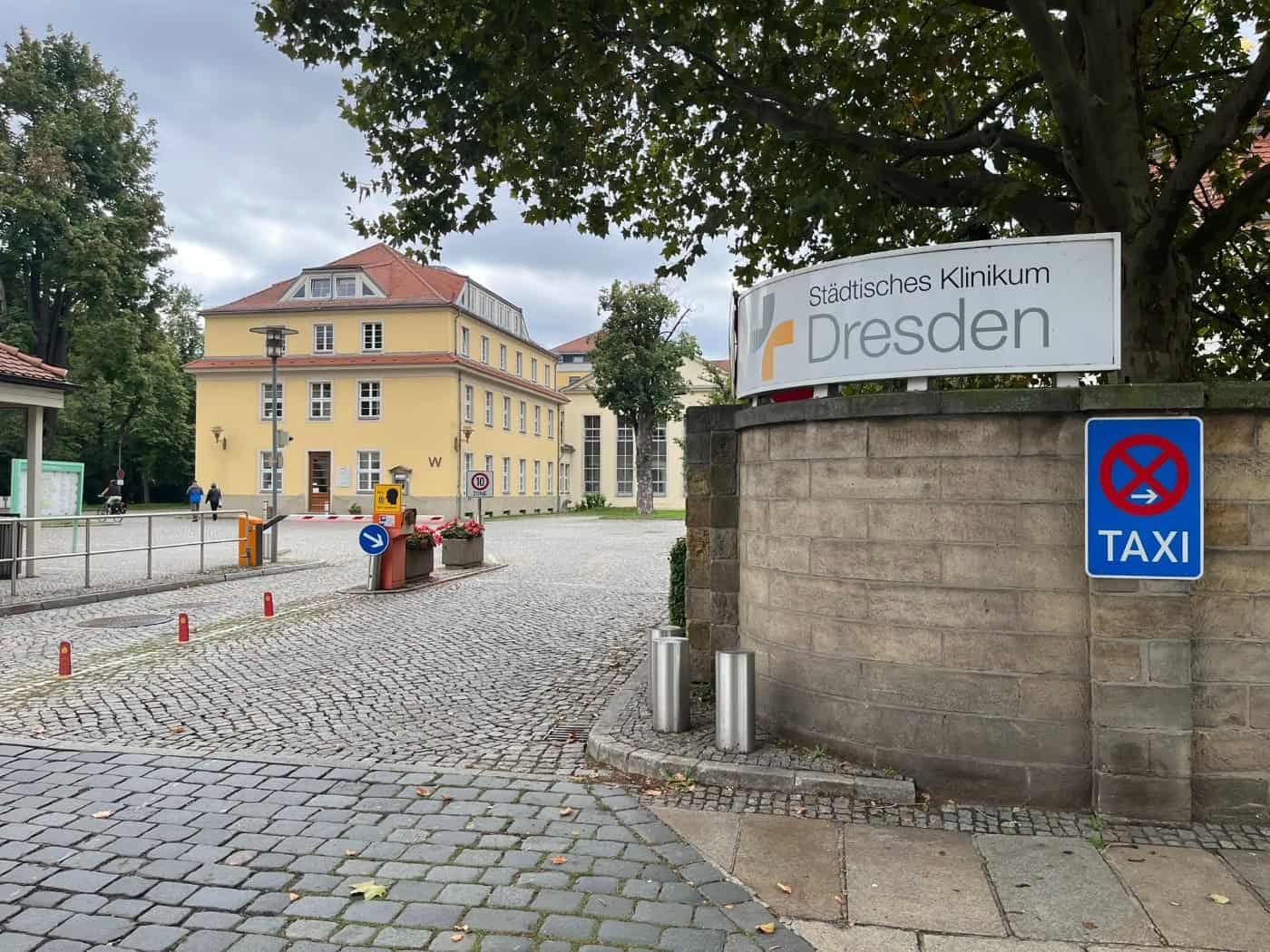 Gepflasterte Einfahrt zum Städtischen Klinikum Dresden mit Schranke: Auf einer Natursteinmauer im Vordergrund steht ein großes Schild mit dem Klinikslogan. Im Hintergrund befindet sich ein dreigeschossiges gelbes Gebäude mit Walmdach.
