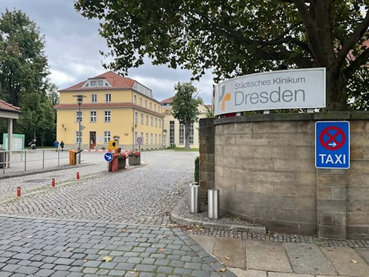 Gepflasterte Einfahrt zum Städtischen Klinikum Dresden mit Schranke: Auf einer Natursteinmauer im Vordergrund steht ein großes Schild mit dem Klinikslogan. Im Hintergrund befindet sich ein dreigeschossiges gelbes Gebäude mit Walmdach.