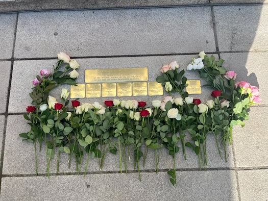 Blick von oben auf sieben im Steinboden verlegte goldene Stolpersteine und einen darüber liegenden Kopfstein. Die Messingtafeln sind umringt von abgelegten roten und weißen Rosen.