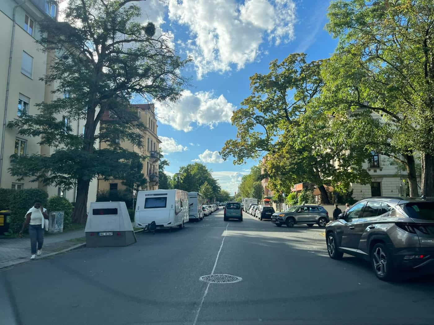 Blick entlang einer Wohnstraße in Dresden mit Bäumen. Auf dem linken Gehweg steht ein grauer, keilförmiger Messanhänger (HWI-VE 167) vor einem geparkten Wohnwagen. Autos fahren auf der Fahrbahn, eine Fußgängerin läuft am Blitzer vorbei.