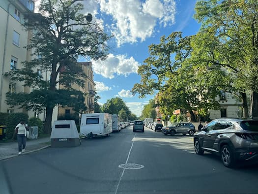 Blick entlang einer Wohnstraße in Dresden mit Bäumen. Auf dem linken Gehweg steht ein grauer, keilförmiger Messanhänger (HWI-VE 167) vor einem geparkten Wohnwagen. Autos fahren auf der Fahrbahn, eine Fußgängerin läuft am Blitzer vorbei.