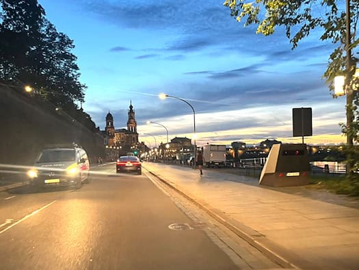 Abendlicher Blick aus der Windschutzscheibe auf das Terrassenufer in Dresden. Rechts auf dem Gehweg steht ein grauer Messanhänger (HWI-VE 159), links fährt ein Polizeiauto. Im Hintergrund leuchten Straßenlaternen und die Türme der historischen Altstadt.