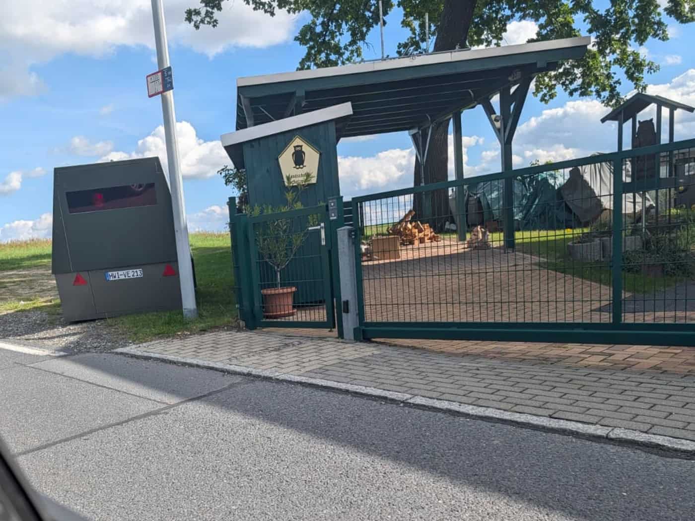 Blick aus dem Autofester auf den gepanzerten Messanhänger HWI-VE 213, der neben einem grünen Hoftor und einer Straßenlaterne auf dem Randstreifen einer schmalen Asphaltstraße steht. Dahinter erstrecken sich Felder.