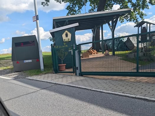 Blick aus dem Autofester auf den gepanzerten Messanhänger HWI-VE 213, der neben einem grünen Hoftor und einer Straßenlaterne auf dem Randstreifen einer schmalen Asphaltstraße steht. Dahinter erstrecken sich Felder.