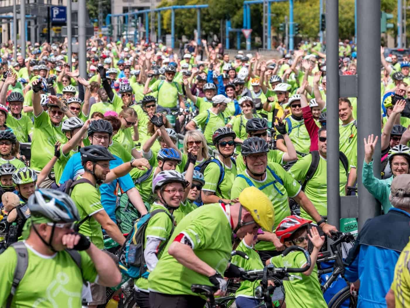 Eine große Menge von Radfahrerinnen und Radfahrern in auffälligen neongrünen T-Shirts und Helmen steht dicht an dicht auf einer Straße in Dresden. Viele lächeln und winken fröhlich in die Kamera.