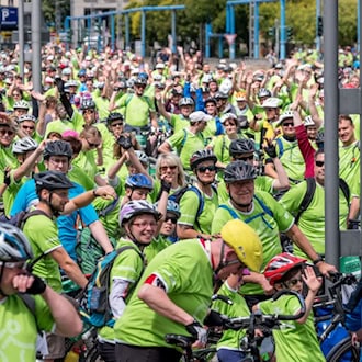Eine große Menge von Radfahrerinnen und Radfahrern in auffälligen neongrünen T-Shirts und Helmen steht dicht an dicht auf einer Straße in Dresden. Viele lächeln und winken fröhlich in die Kamera.