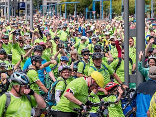Eine große Menge von Radfahrerinnen und Radfahrern in auffälligen neongrünen T-Shirts und Helmen steht dicht an dicht auf einer Straße in Dresden. Viele lächeln und winken fröhlich in die Kamera.