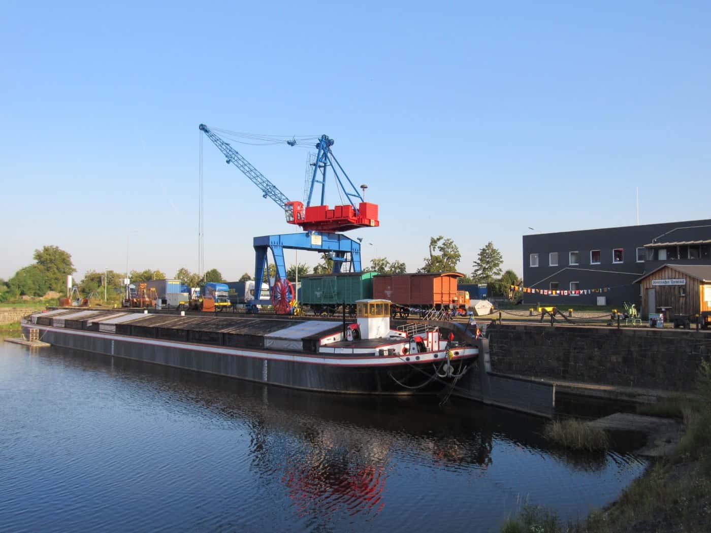 Ein historischer Elbeschleppkahn liegt am Kai eines Hafens. Dahinter steht ein großer, blau-roter Gantry-Kran. Am Kai Güterwagen und ein graues Industriegebäude unter klarem blauem Himmel. Ruhiges Wasser spiegelt den Kahn.