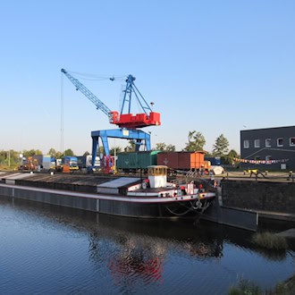 Ein historischer Elbeschleppkahn liegt am Kai eines Hafens. Dahinter steht ein großer, blau-roter Gantry-Kran. Am Kai Güterwagen und ein graues Industriegebäude unter klarem blauem Himmel. Ruhiges Wasser spiegelt den Kahn.