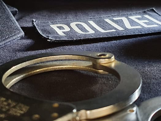 Nahaufnahme von metallenen Handschellen, die auf einer dunklen Polizei-Uniform mit dem Schriftzug POLIZEI liegen. Im Hintergrund befinden sich gestapelte, hellblaue Diensthemden.