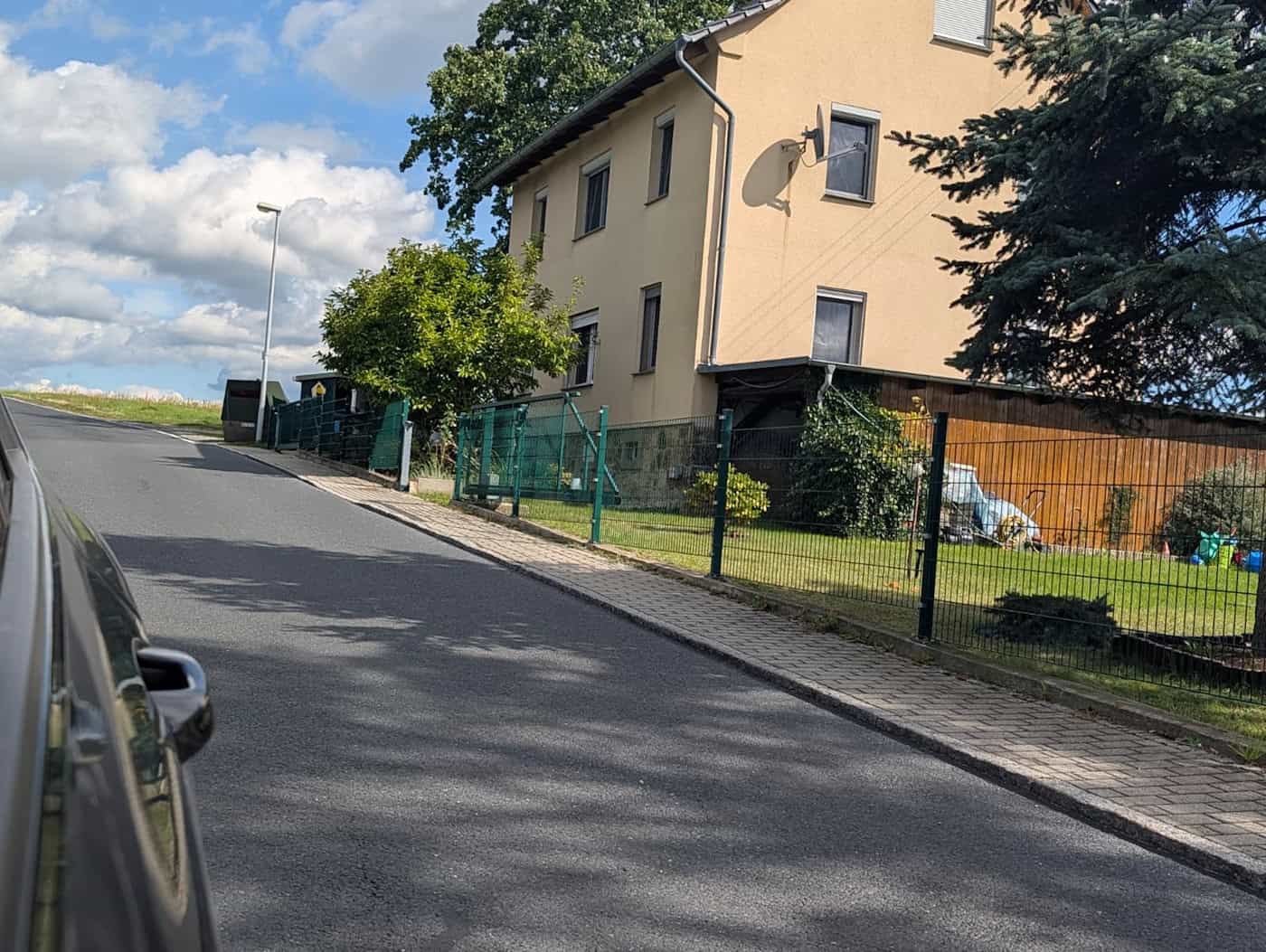 Schmale Grünberger Straße in Hermsdorf mit Wohnhäusern und Blitzer am Straßenrand Blick entlang einer steil ansteigenden, schmalen Wohnstraße. Rechts stehen Einfamilienhäuser mit Zäunen und Gärten, weiter oben am Hügel ist am linken Straßenrand ein dunkler Messanhänger platziert.