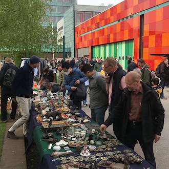 Besucher stöbern an einem langen Tisch voller Trödel, Messing und Antiquitäten. Im Hintergrund stehen das Hochhaus vom Haus der Presse Dresden und ein Gebäude mit bunt rot-orange gemusterter Fassade.