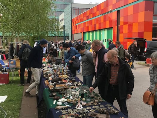 Besucher stöbern an einem langen Tisch voller Trödel, Messing und Antiquitäten. Im Hintergrund stehen das Hochhaus vom Haus der Presse Dresden und ein Gebäude mit bunt rot-orange gemusterter Fassade.
