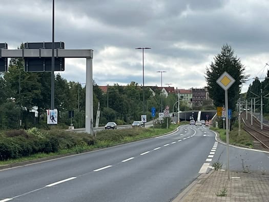 Eine mehrspurige Asphaltstraße führt geschwungen auf die Einfahrt des Bramschstraßen-Tunnels zu. Am Straßenrand stehen Verkehrsschilder, Straßenlaternen und Bäume, rechts verlaufen Straßenbahnschienen. Im Hintergrund Autos vor der Tunneleinfahrt unter grauem Himmel.