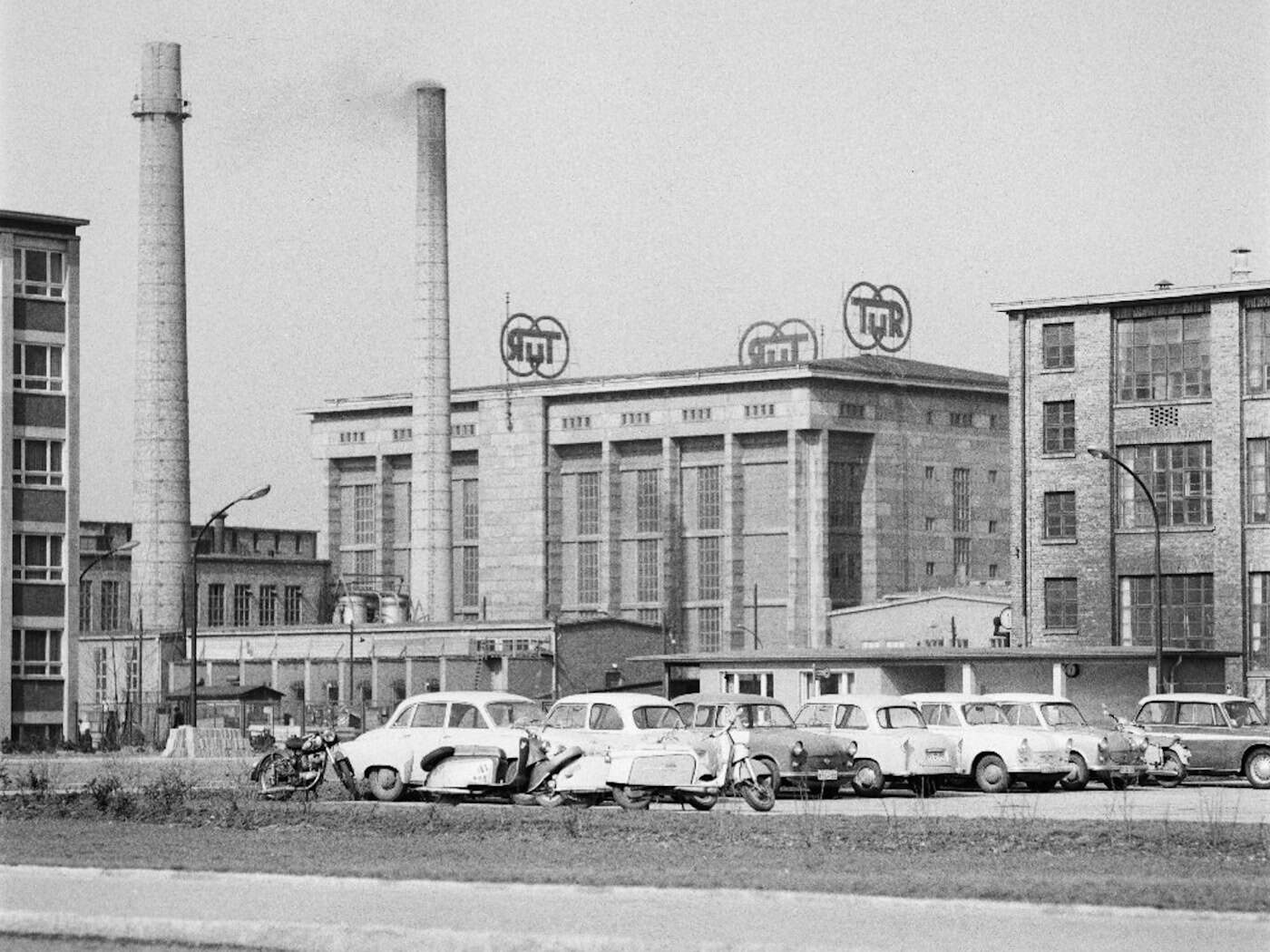 Historische Aufnahme des VEB Transformatoren- und Röntgenwerk (TuR) Dresden Schwarz-Weiß-Aufnahme des VEB Transformatoren- und Röntgenwerk in Dresden mit TuR-Logo auf dem Dach, im Vordergrund rauchende Schornsteine sowie parkende DDR-Oldtimer und Mopeds.