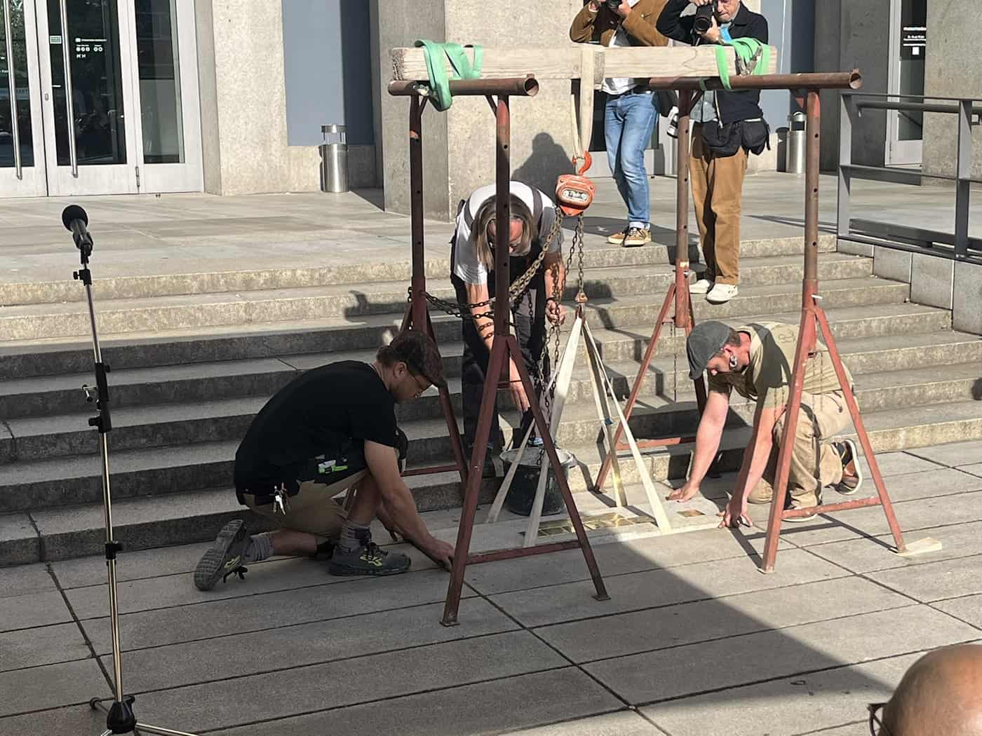 Handwerker knien auf dem Vorplatz vor den Museumsstufen und passen mit Hilfsgerüsten goldene Stolpersteine in die Gehwegplatten ein. Fotografen beobachten die Arbeiten.