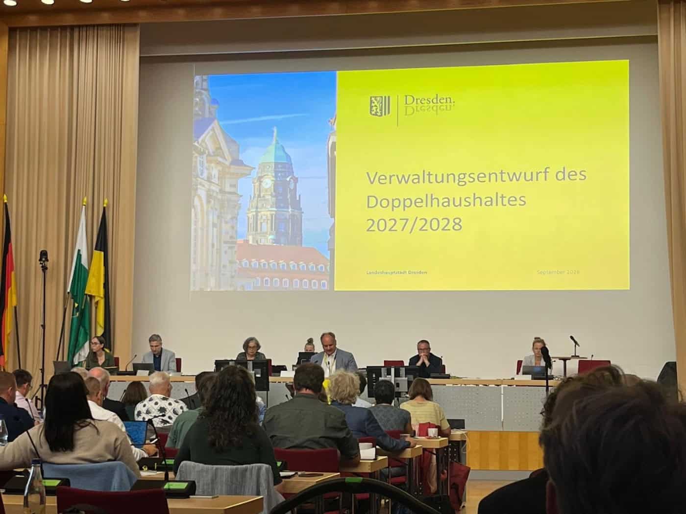 Blick in den Plenarsaal des Dresdner Stadtrats: Auf einer großen Leinwand ist der Schriftzug „Verwaltungsentwurf des Doppelhaushaltes 2027/2028“ zu sehen. Davor sitzen Stadtpolitiker an einem Podiumstisch, im Vordergrund blickt man auf das Publikum.
