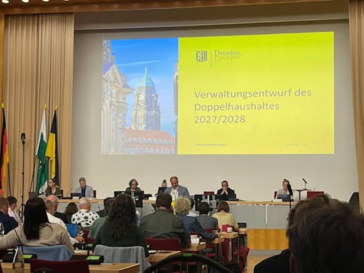 Blick in den Plenarsaal des Dresdner Stadtrats: Auf einer großen Leinwand ist der Schriftzug „Verwaltungsentwurf des Doppelhaushaltes 2027/2028“ zu sehen. Davor sitzen Stadtpolitiker an einem Podiumstisch, im Vordergrund blickt man auf das Publikum.