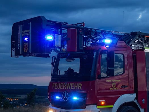 Ein rotes Drehleiter-Feuerwehrfahrzeug steht bei Dämmerung mit eingeschaltetem Blaulicht und leuchtenden Scheinwerfern auf einem Platz. Die Drehleiter ist eingefahren, im Hintergrund zeigt sich eine Landschaft unter abendlichem Himmel.
