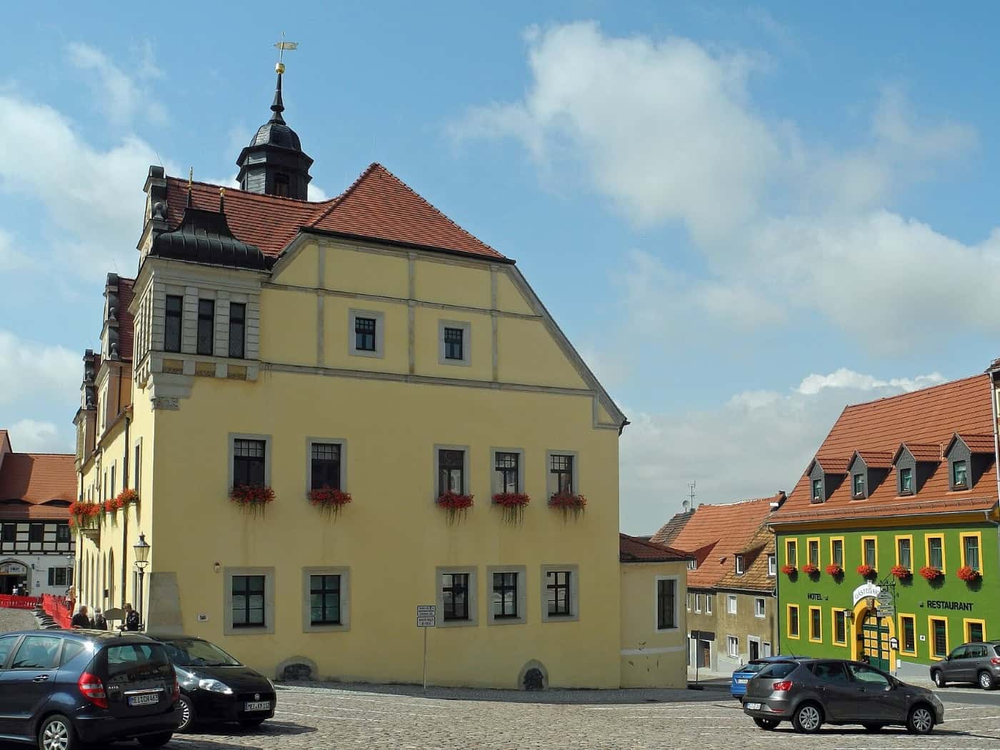 Gelbes historisches Rathaus mit Turmhaube und roten Blumenkästen am Markt in Lommatzsch, davor parkende Autos