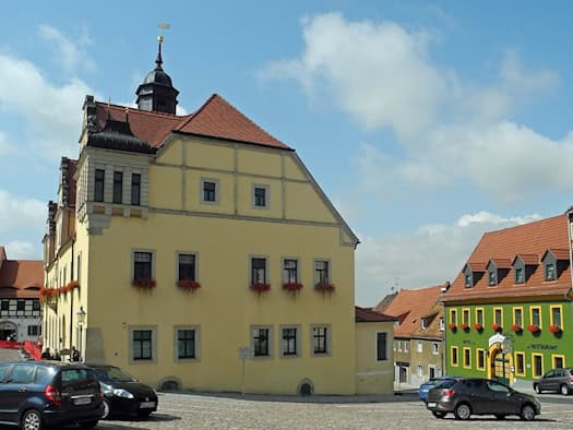 Gelbes historisches Rathaus mit Turmhaube und roten Blumenkästen am Markt in Lommatzsch, davor parkende Autos