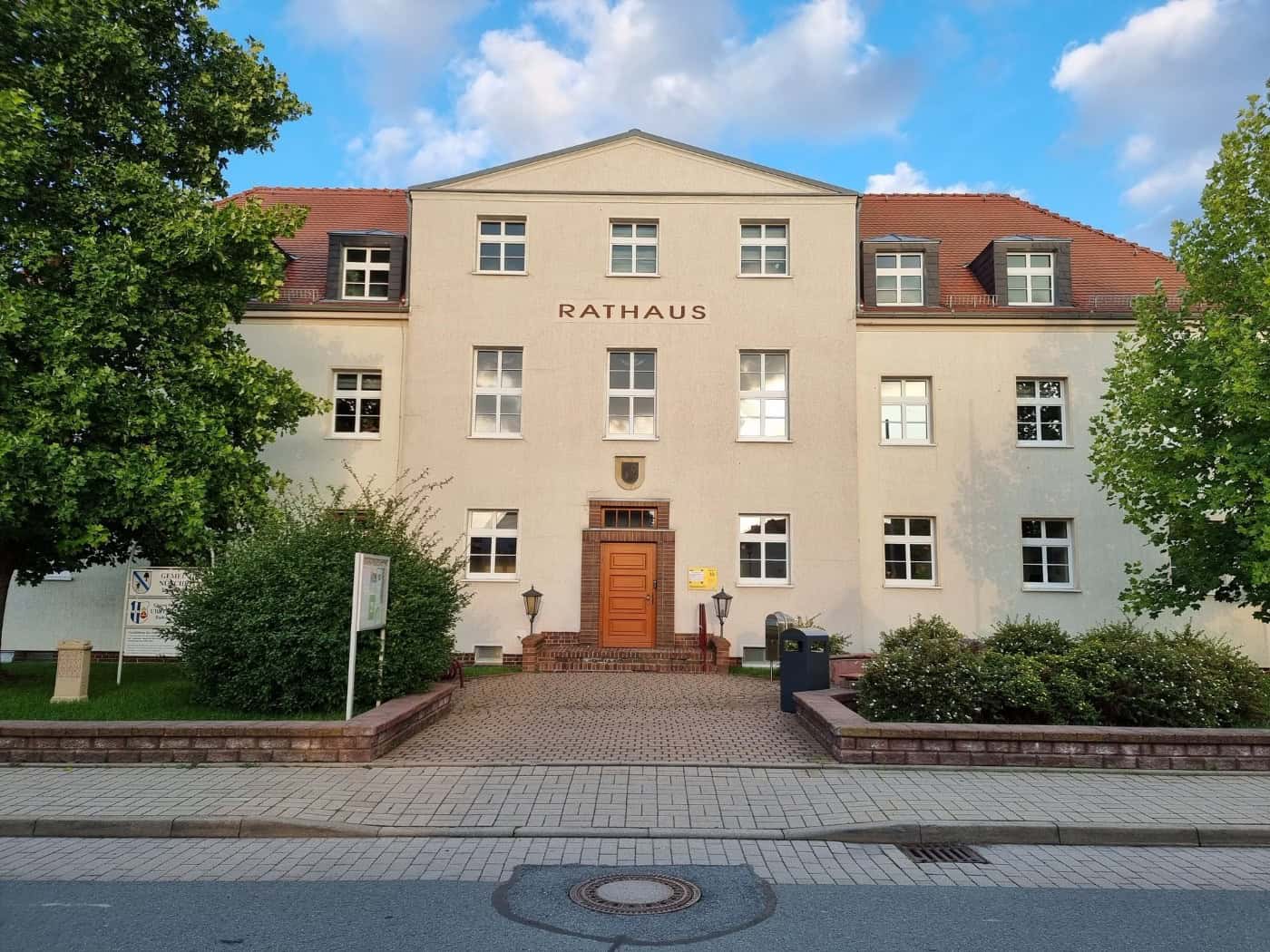 Zweistöckiges helles Rathausgebäude mit Schriftzug Rathaus und Holztür, davor ein gepflasterter Weg und Bäume