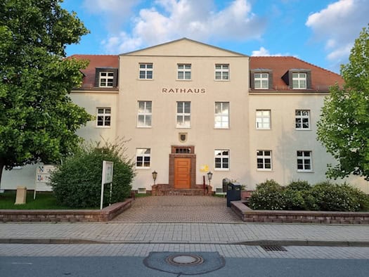 Zweistöckiges helles Rathausgebäude mit Schriftzug Rathaus und Holztür, davor ein gepflasterter Weg und Bäume