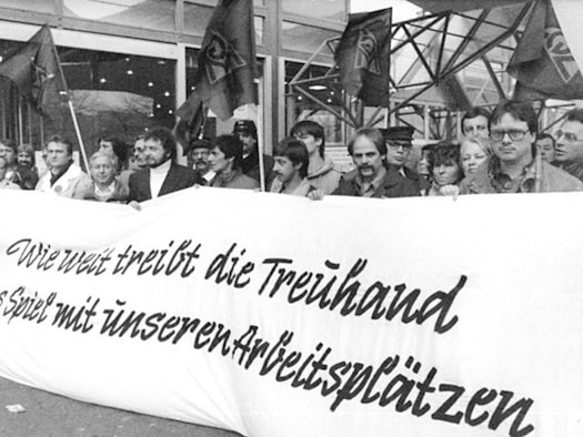Schwarz-Weiß-Foto: Demonstranten mit Fahnen halten ein Transparent mit der Aufschrift Wie weit treibt die Treuhand das Spiel mit unseren Arbeitsplätzen