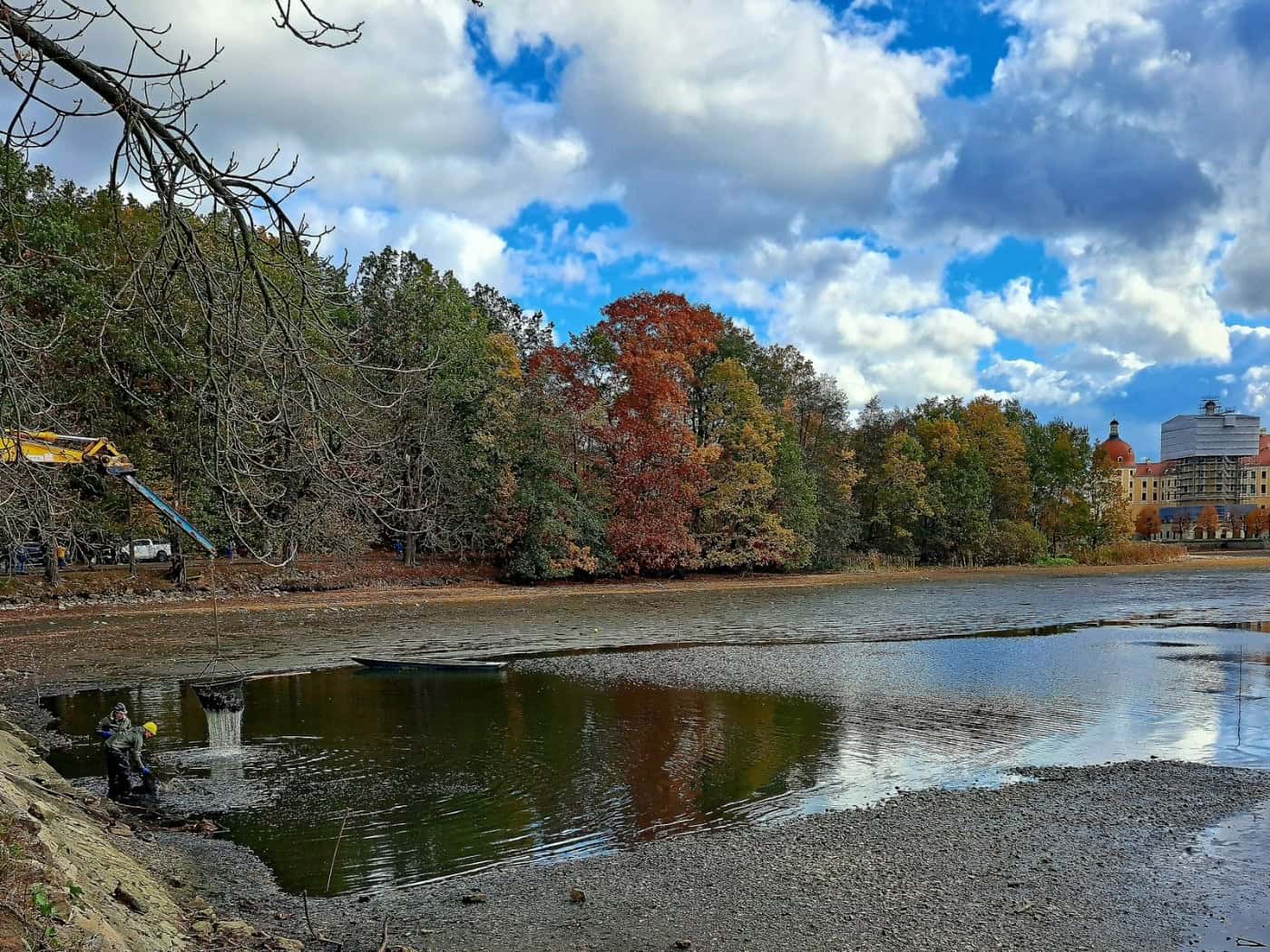 Teilweise abgelassener Teich vor Herbstwald, rechts im Hintergrund Schloss Moritzburg, links ein Kranarm und Arbeiter am Ufer