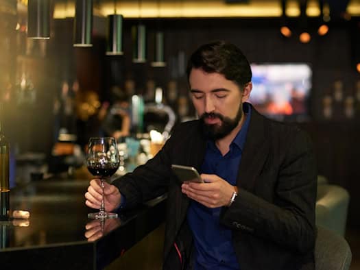 Mann im Sakko sitzt abends in einer Bar mit einem Glas Rotwein und einem Smartphone in der Hand