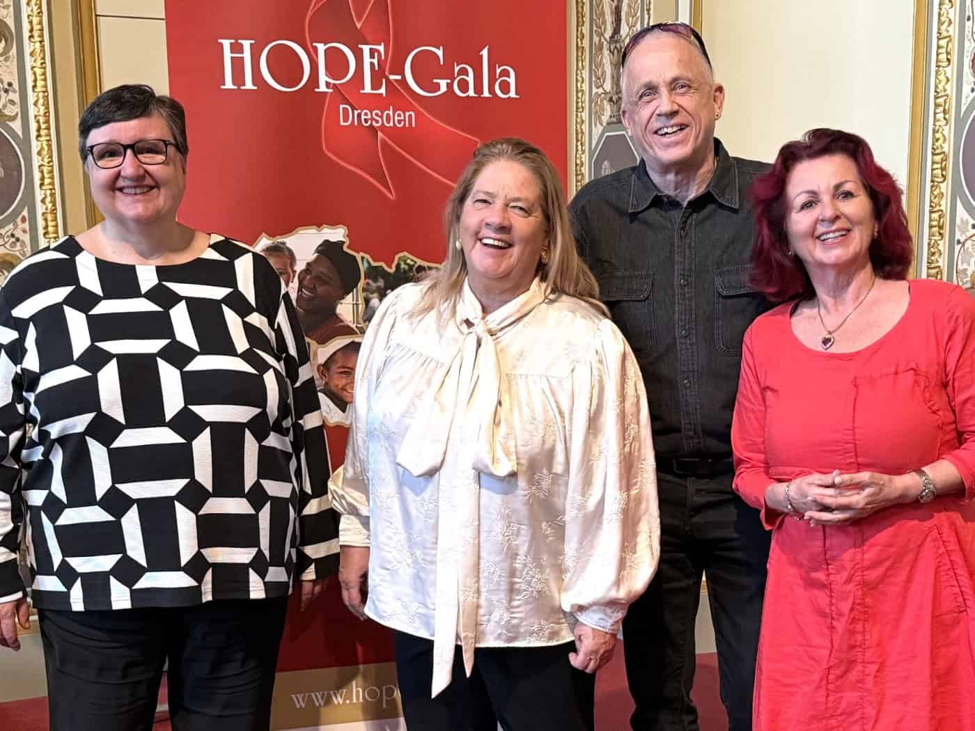 Vier lächelnde Personen stehen nebeneinander vor einem roten Banner der HOPE-Gala Dresden: Dr. Maria Decker in gemustertem Oberteil, Kathy Kelly in weißer Bluse, Stefan Hippler in dunklem Hemd und Viola Klein in einem knalligen pinken Kleid.