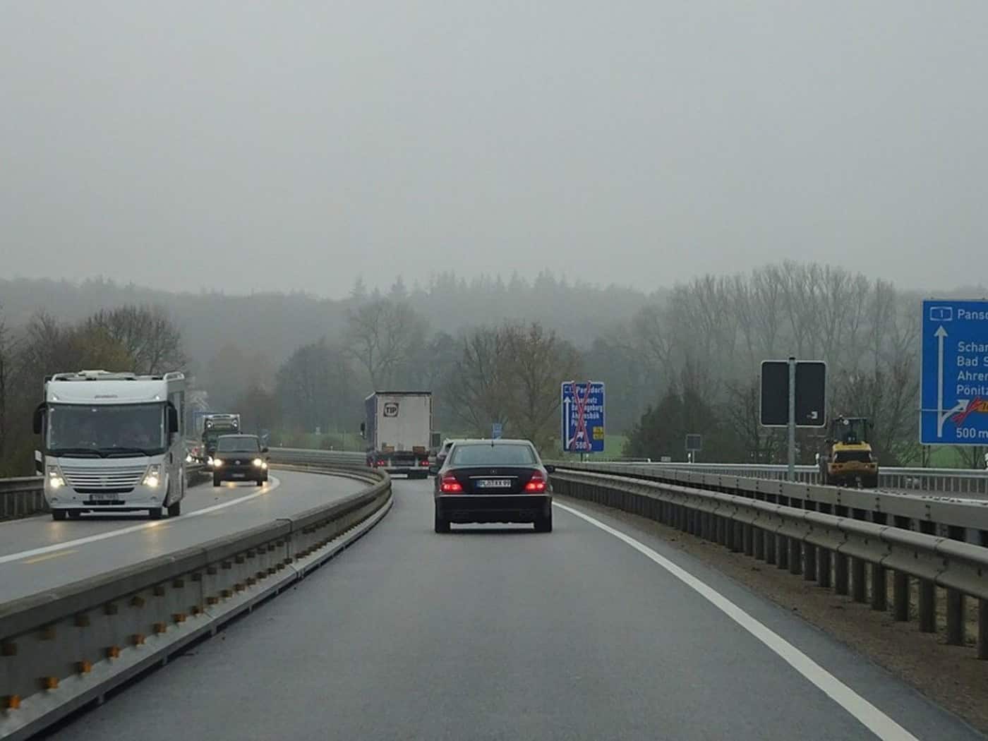 Autos und Lastwagen fahren auf einer Autobahn im Nebel, rechts ein blaues Ausfahrtsschild und eine Walze am Fahrbahnrand.
