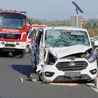 Weißer Ford-Transporter mit zerstörter Front und zersplitterter Frontscheibe auf der A4, dahinter ein Löschfahrzeug der Feuerwehr und Einsatzkräfte, im Hintergrund Stau und Windräder