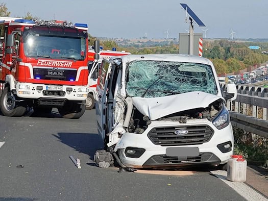 Weißer Ford-Transporter mit zerstörter Front und zersplitterter Frontscheibe auf der A4, dahinter ein Löschfahrzeug der Feuerwehr und Einsatzkräfte, im Hintergrund Stau und Windräder