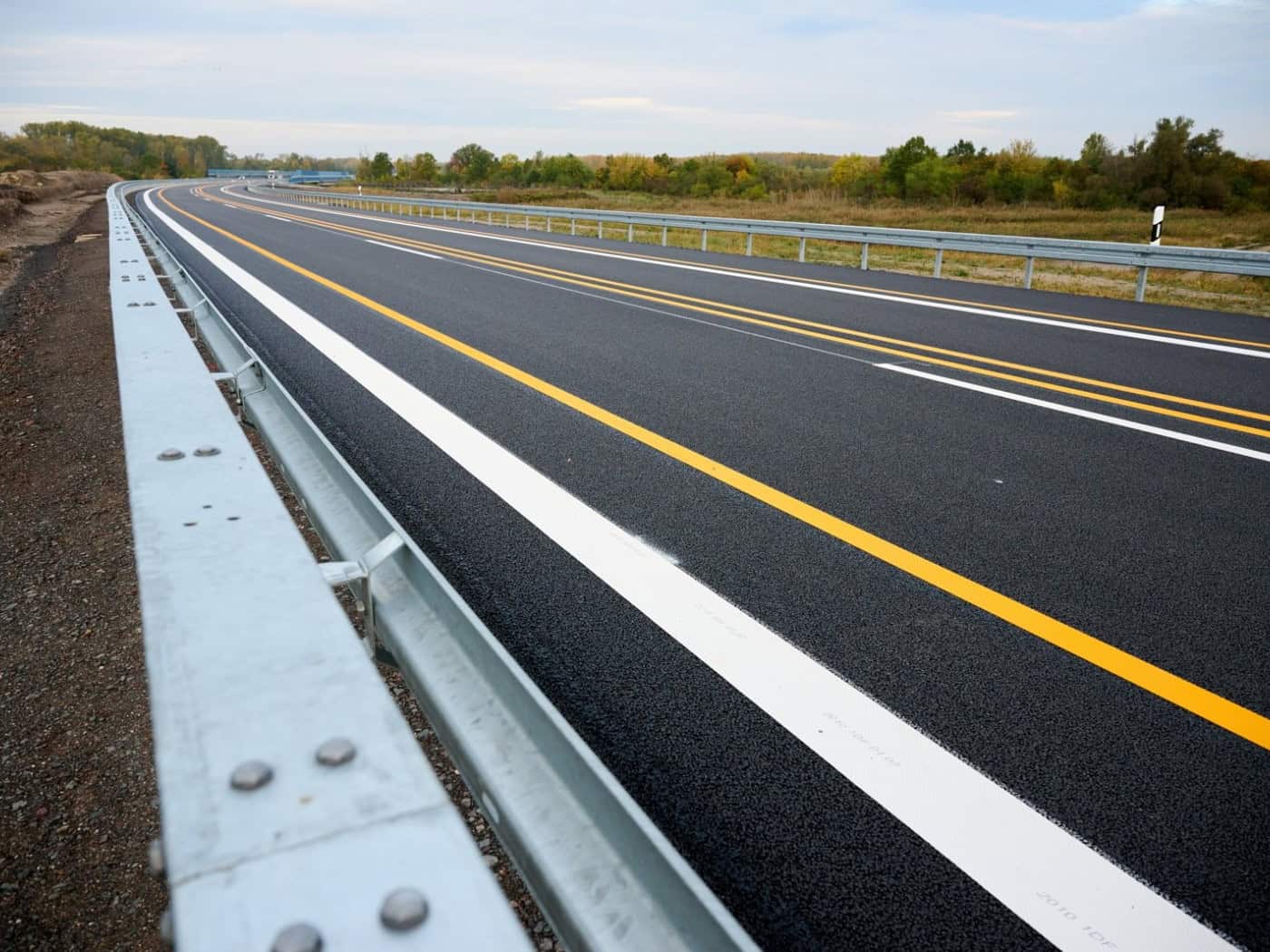 Neue, leere Autobahnfahrbahn mit gelben Baustellenmarkierungen und Leitplanke an einem bewölkten Herbsttag