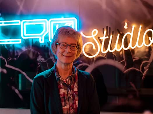 Porträt von Dr. Alexandra-Kathrin Stanislaw-Kemenah mit Brille und kariertem Hemd, die lächelnd vor einer blau-orange leuchtenden Neon-Aufschrift („C90 Studio“) steht.